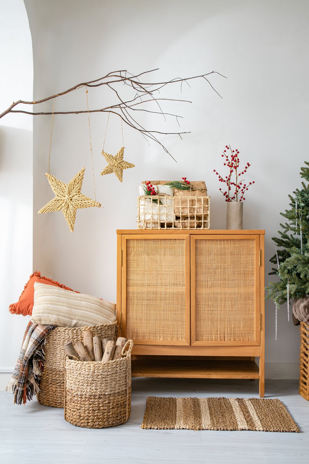 Handwoven natural fiber Christmas stars hanging from a bare branch above a rattan cabinet, with water hyacinth baskets and a jute rug